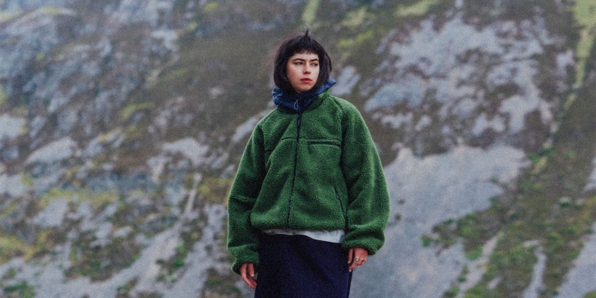 Gramicci Automne-Hiver 2026 : veste polaire verte sur une plage de galets face à la montagne