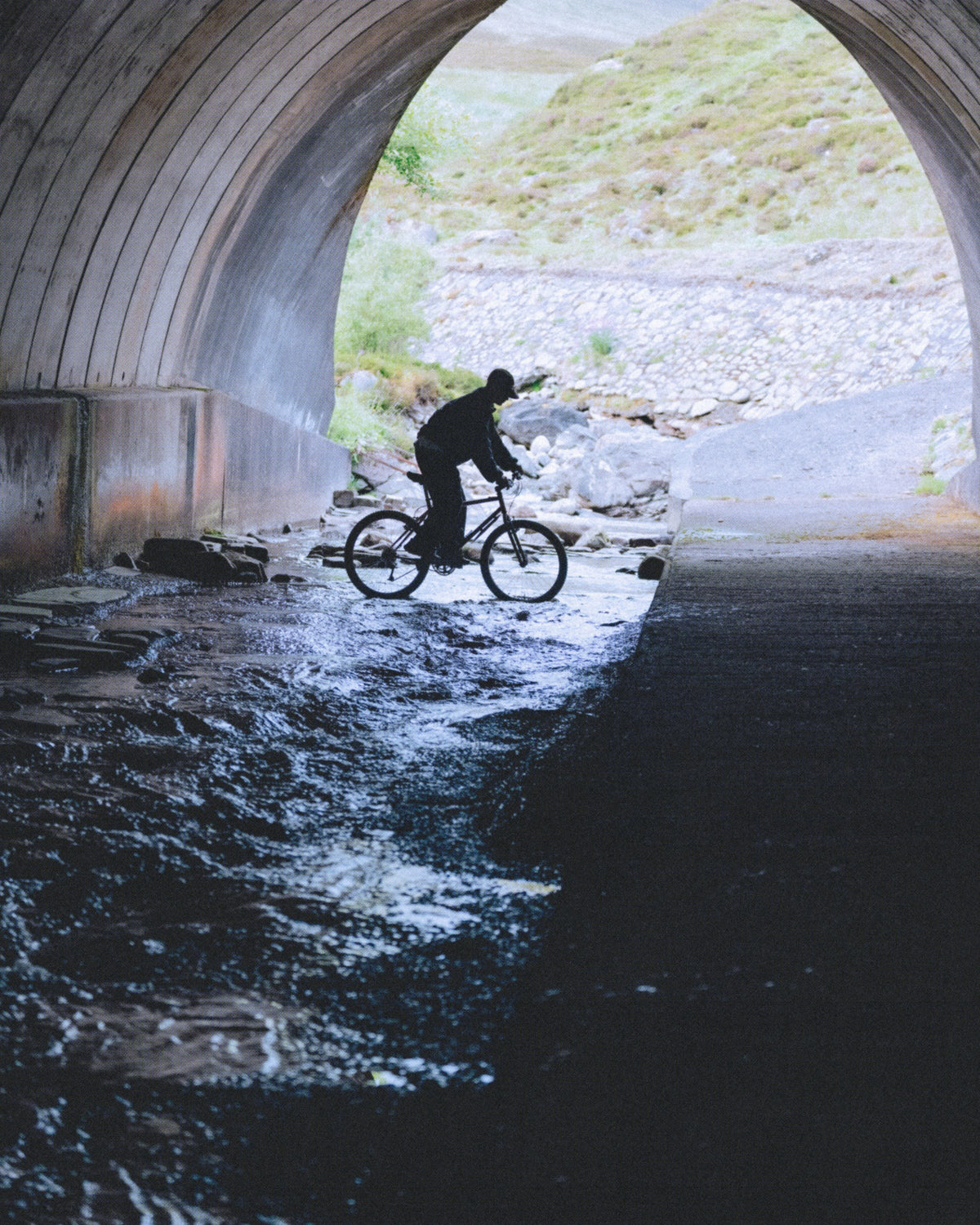 Campagne Gramicci Automne-Hiver 2026 — cycliste traversant l’eau sous un pont en arche